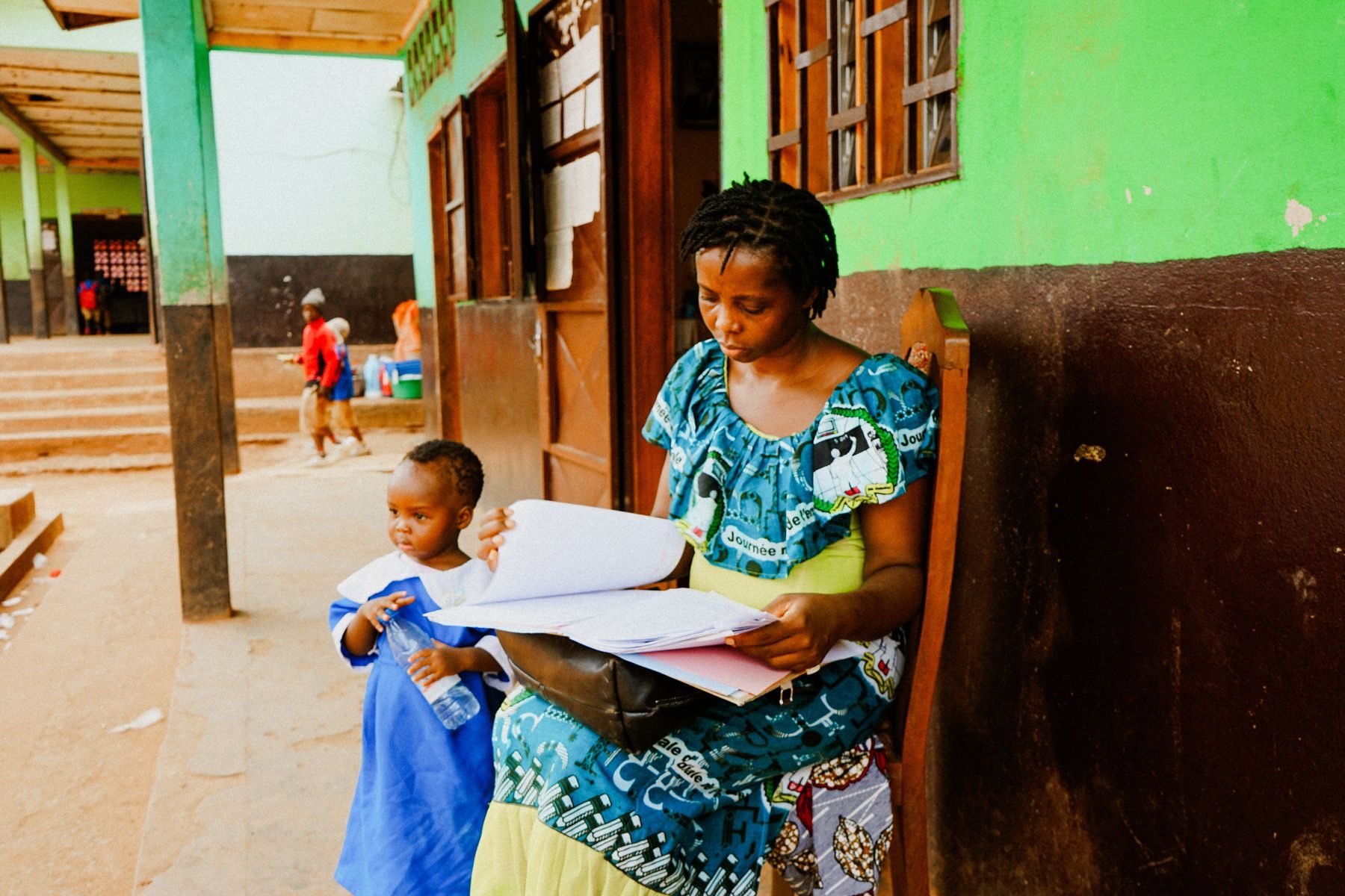A mother reads a school folder at the gate while her child waits beside her