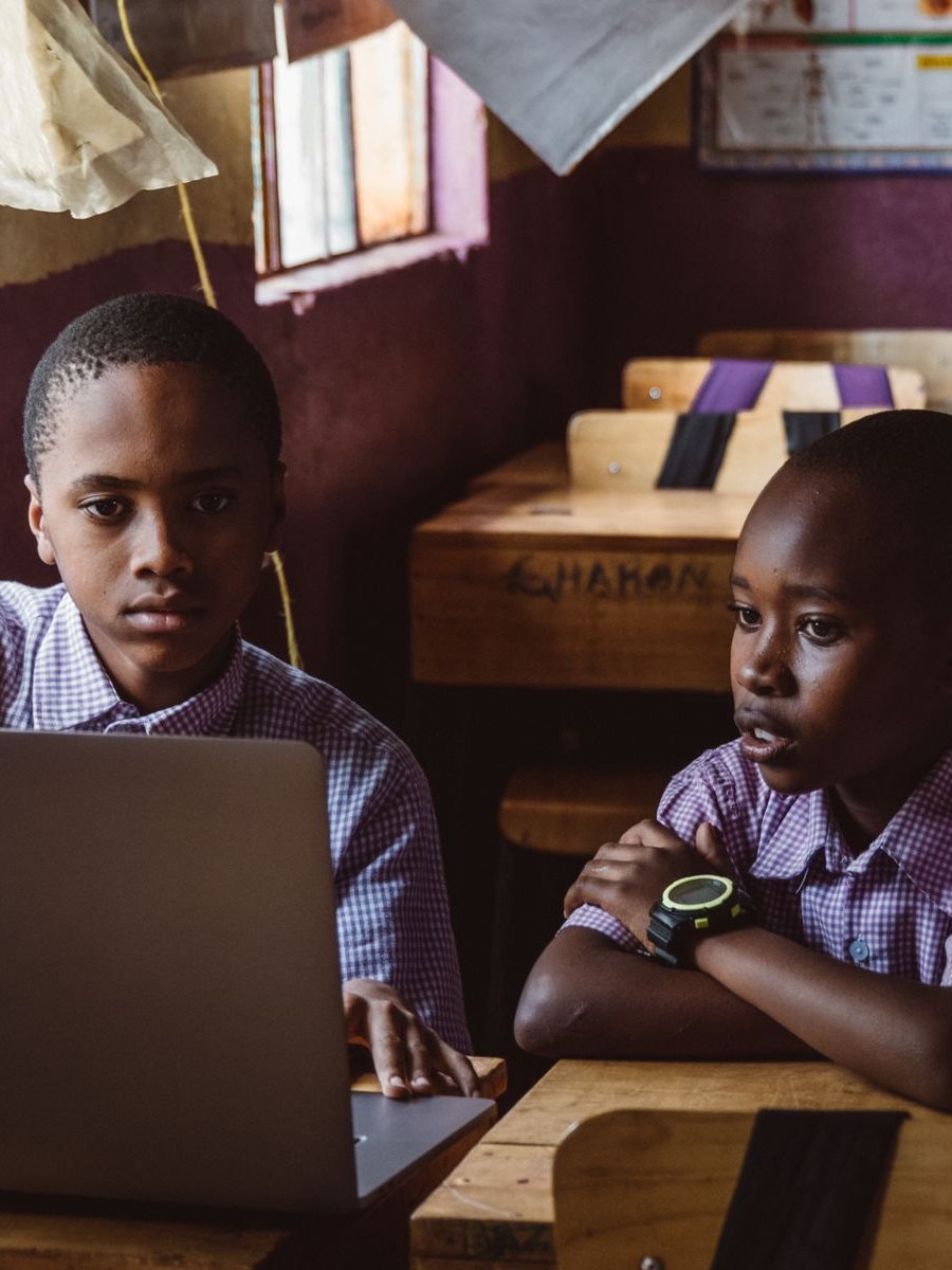 Two boys in school uniform share a laptop at a wooden desk in an African classroom, one looking at the screen and the other listening