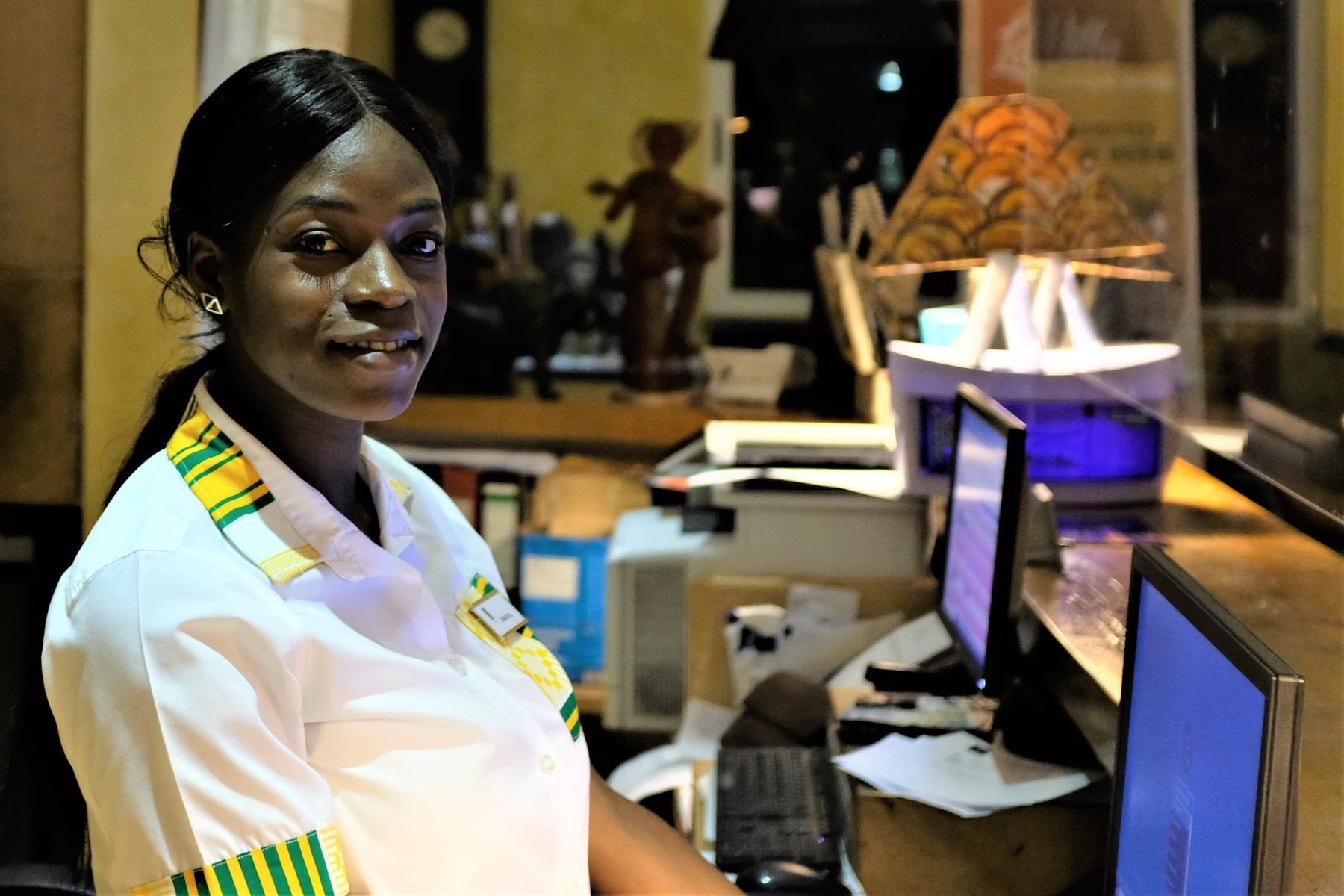 A smiling Ghanaian hotel receptionist in a white uniform with kente trim sits at the front desk beside her monitors in a hotel in Accra, Gha