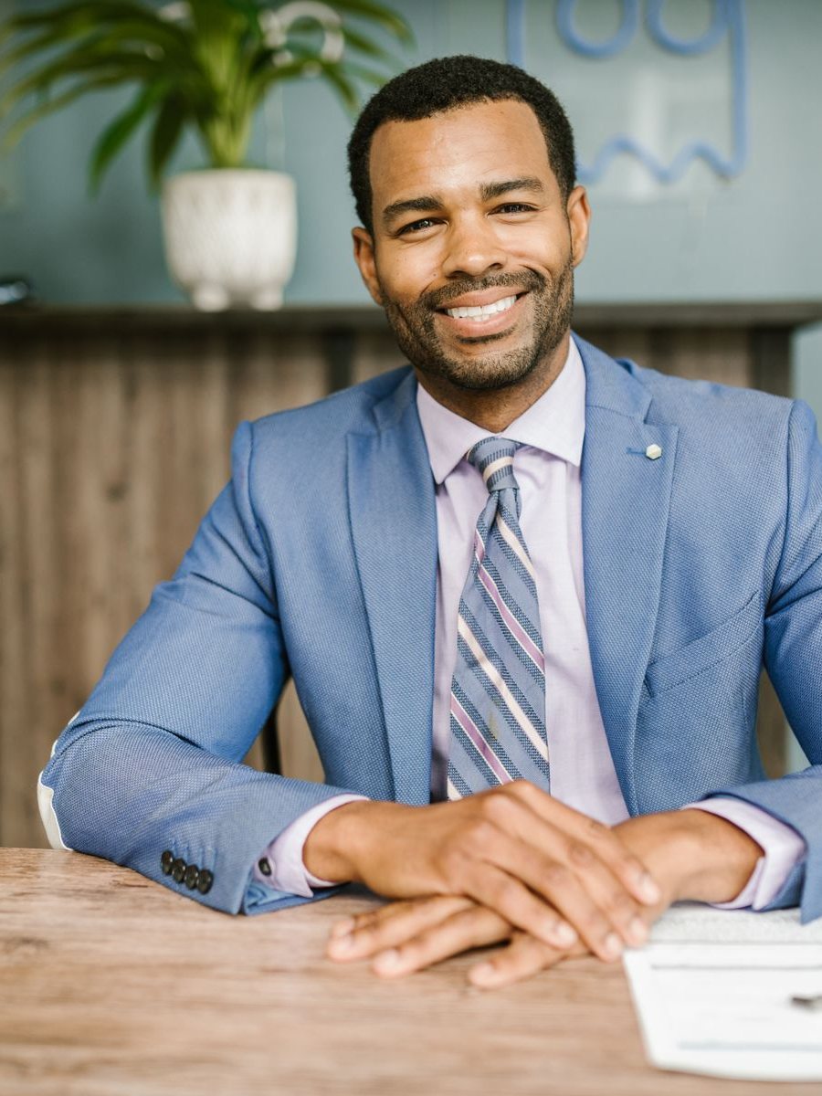 A smiling Black insurance agent in a blue suit and tie seated at a wooden desk with policy documents and a pen in front of him, plants and n